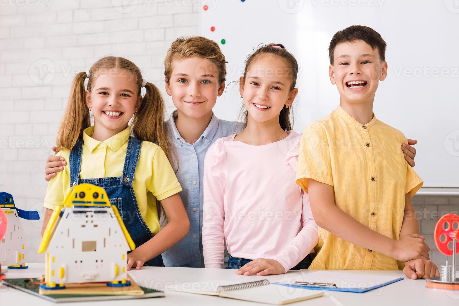 A group of four students are working on a robotics project in a classroom. They are smiling and appear to be enjoying themselves. There are various robot models on the table in front of them. photo