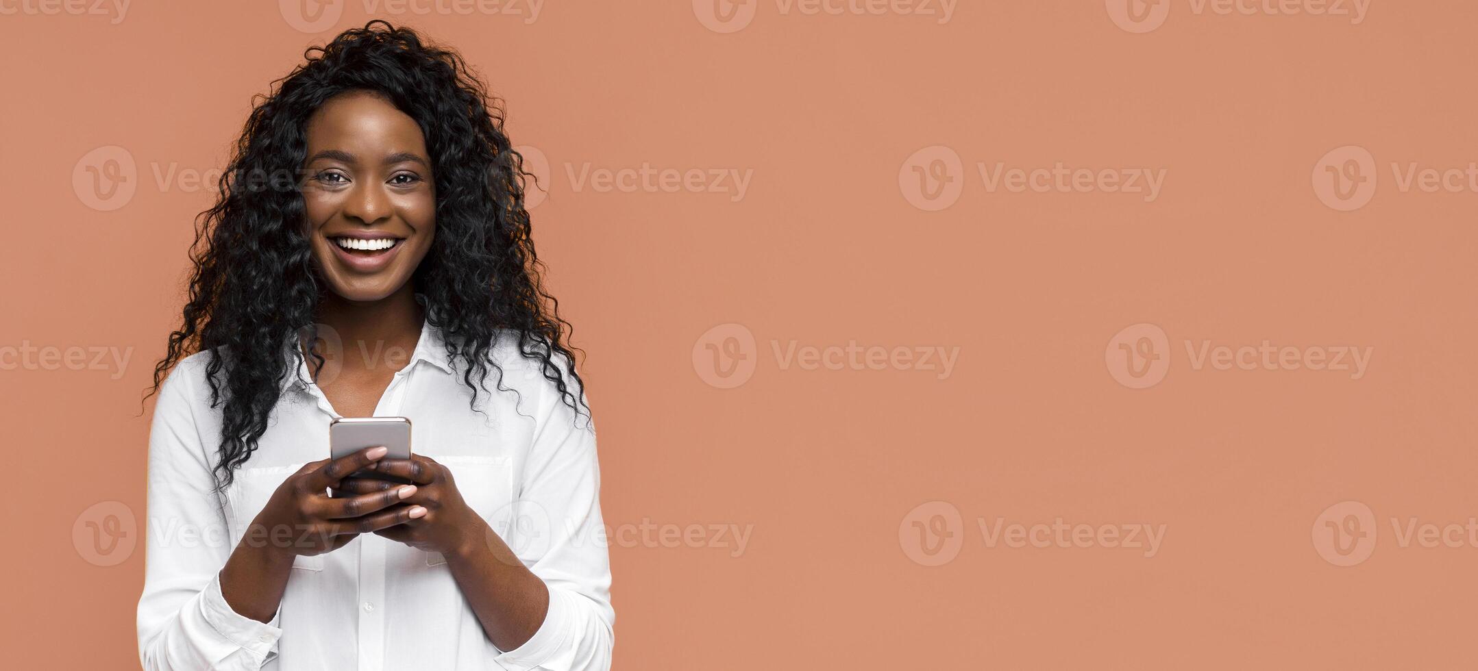 A young Black woman with curly hair smiles while holding a smartphone in her hands. She is dressed in a white button-down shirt and stands in front of a peach background. photo