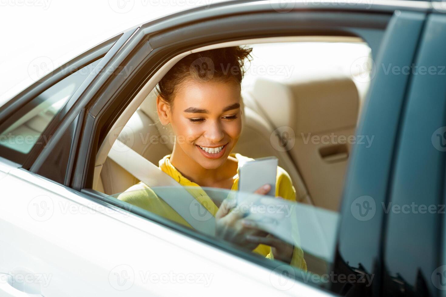 A young woman with dark hair and a bright smile sits in the backseat of a car on a sunny day. She is using her smartphone, presumably scrolling through social media or texting a friend. photo