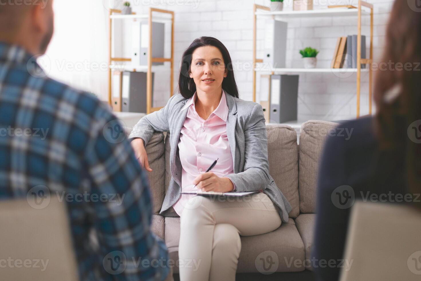 A therapist sits on a couch in an office, listening intently to a client who sits across from her. She is taking notes on a notepad and appears to be focused on the clients words. photo