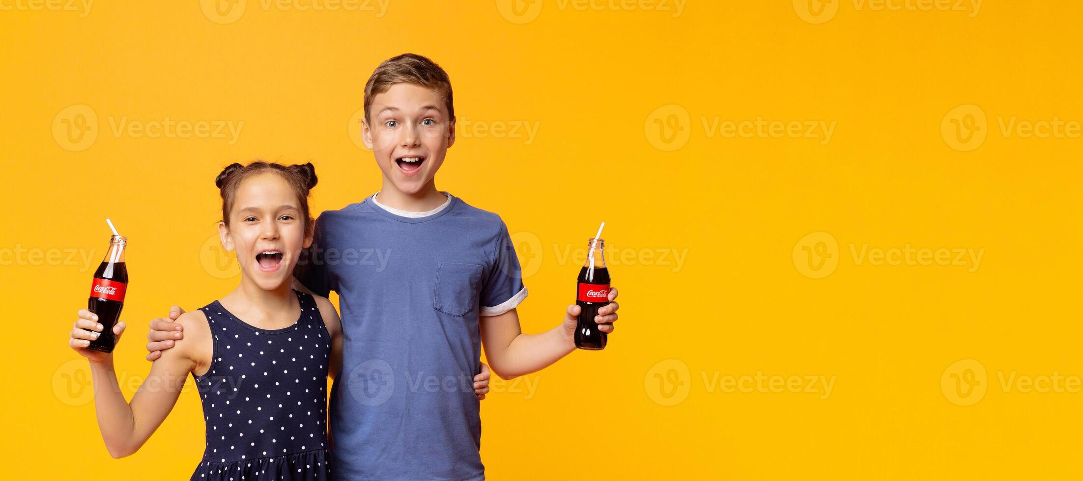 KHARKIV, UKRAINE - April 03. 2019. Happy children drinking Coca-Cola Classic with straws on orange background. The drink is produced and manufactured by The Coca-Cola Company photo