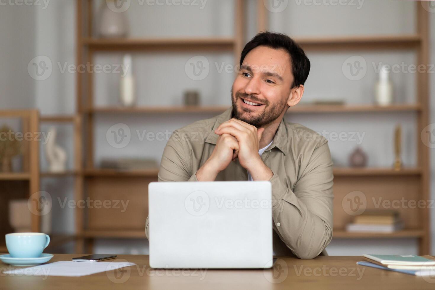 A man is seated at a desk, focused on his laptop screen. He seems engrossed in his work, possibly typing or researching. The desk is cluttered with papers and office supplies. photo