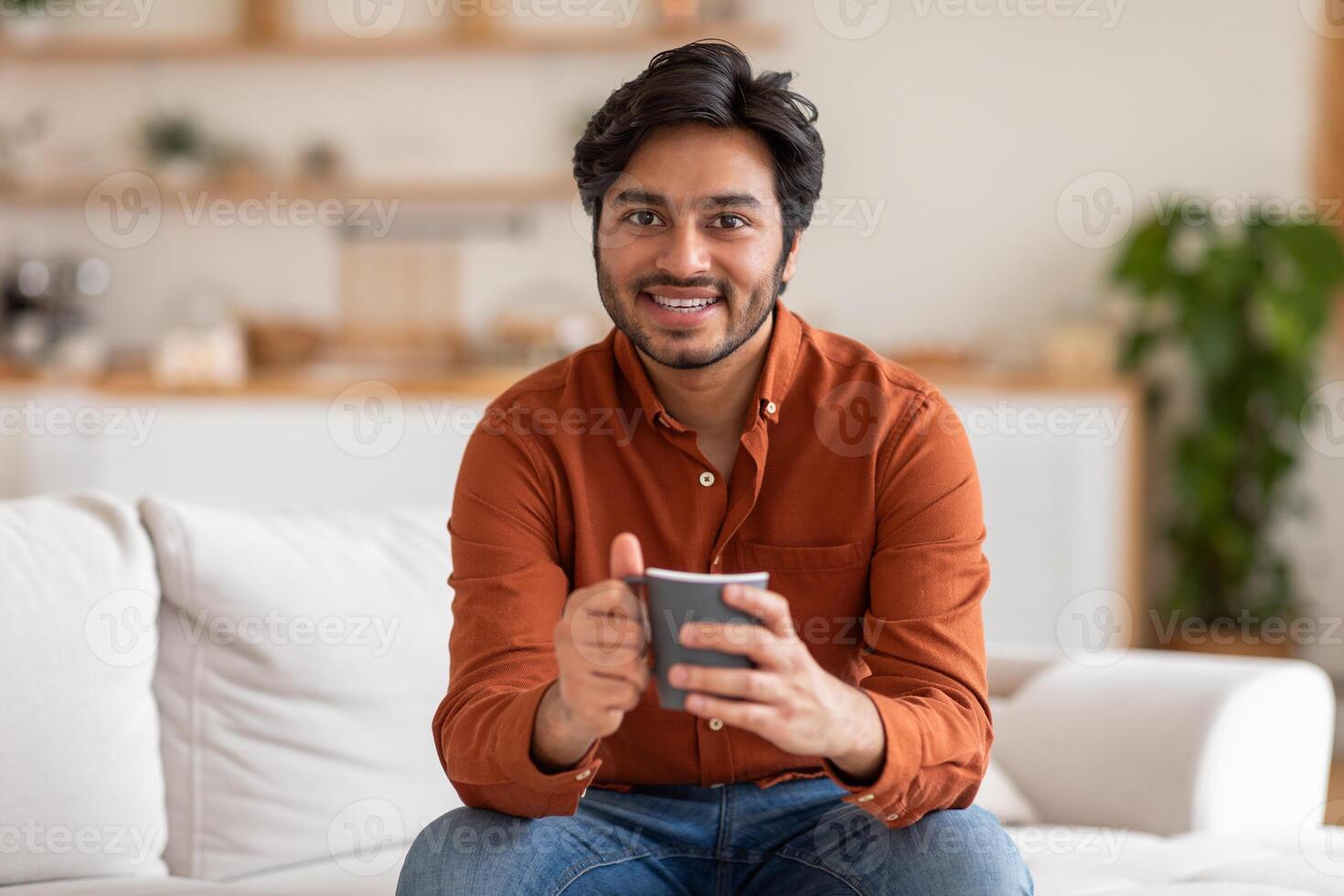 Arab man in an orange shirt sits on a white couch in a living room, smiling and holding a dark gray mug. He appears to be enjoying his coffee break. photo