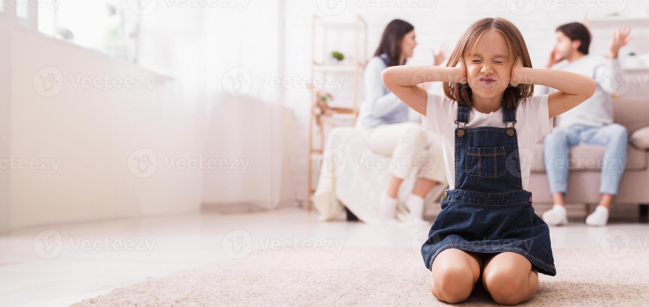 Little girl sitting on floor and covering ears with hands, tired of mom and dad arguing, quarrelling parents on background, selective focus on child photo