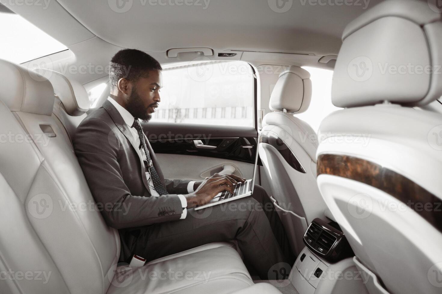 Black businessman working on his laptop in the backseat of a luxury car. He is dressed in a suit and tie, and is focused on his work. The car is spacious and comfortable, with white leather seats photo
