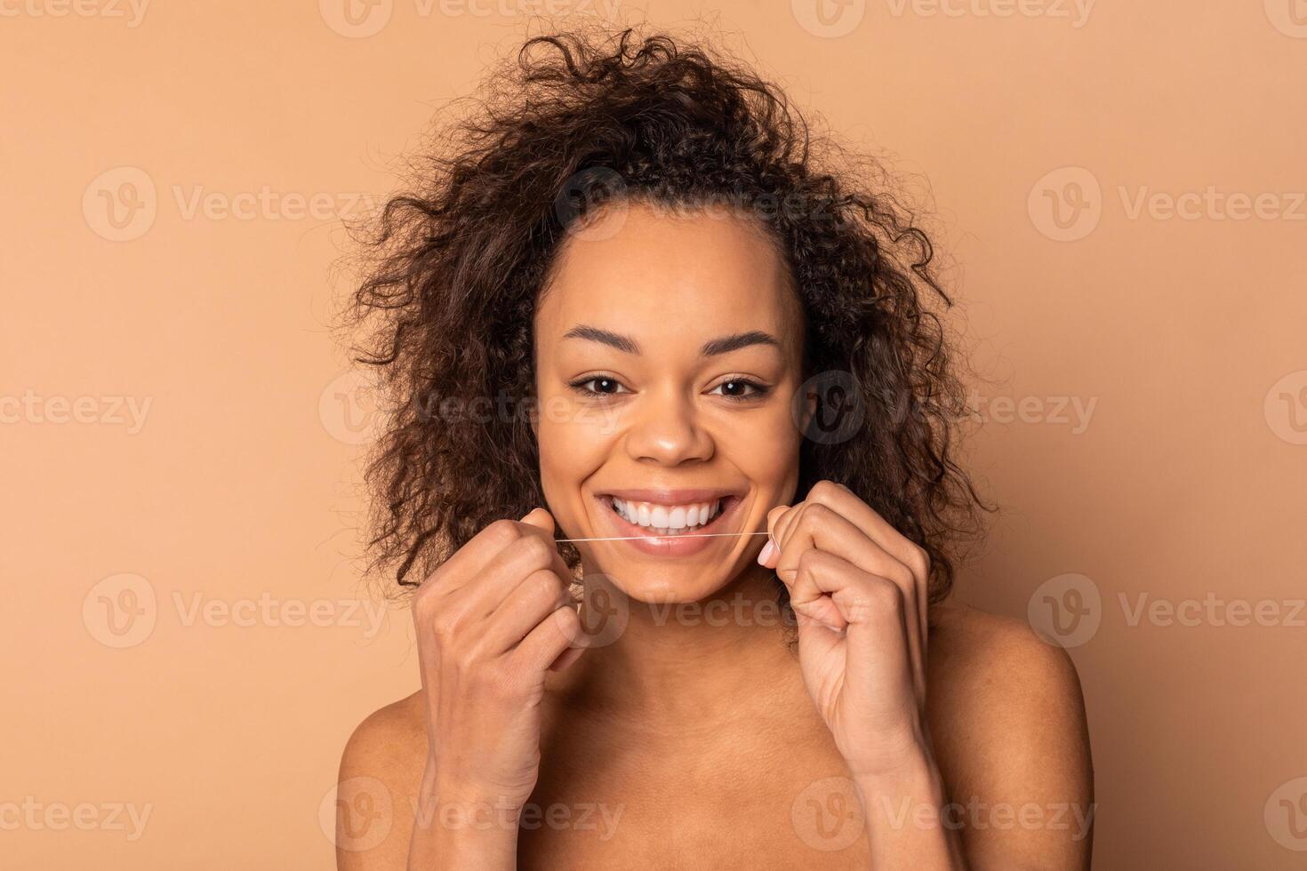 This image shows African American woman with curly brown hair flossing her teeth while smiling. She is wearing no makeup and has a bright white smile. The background is a plain beige color. photo