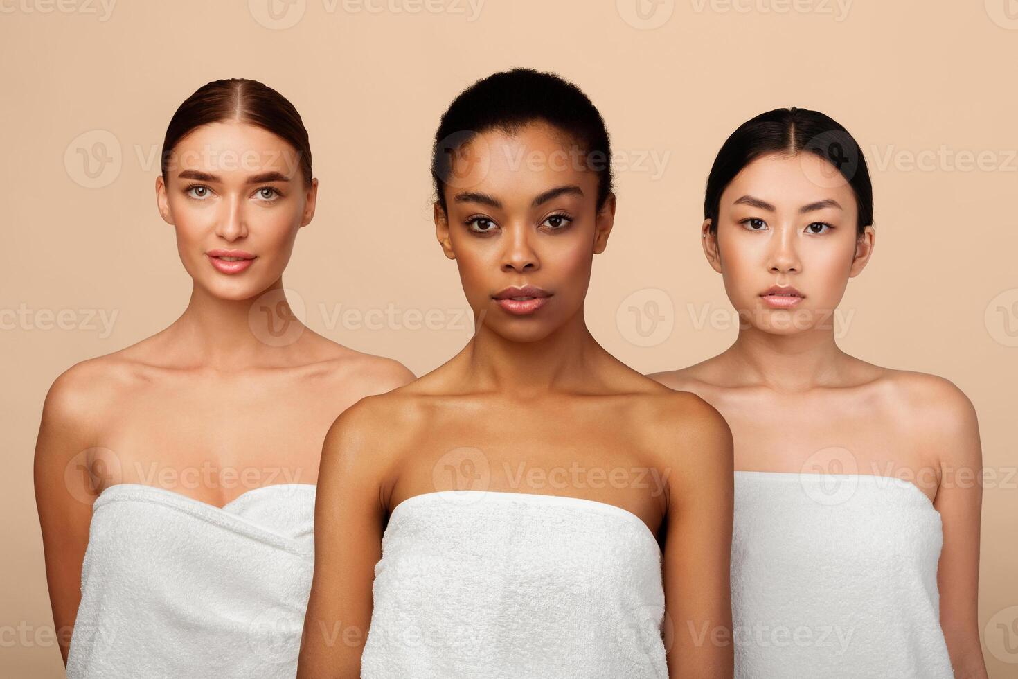 Three Mixed Models Girls Posing Wrapped In Bath Towels Looking At Camera Standing Over Gray Background. Studio Shot photo