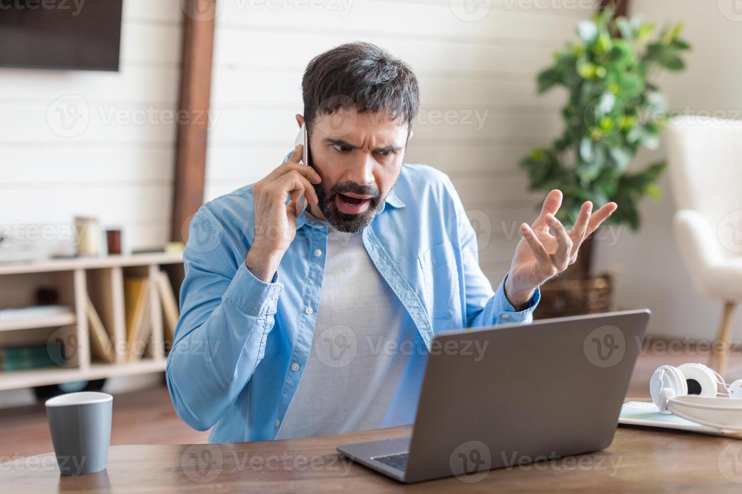 A man appears visibly upset as he argues over the phone, gesturing with his hand. He is seated at a wooden table where his laptop is open, suggesting he is dealing with a work-related issue photo