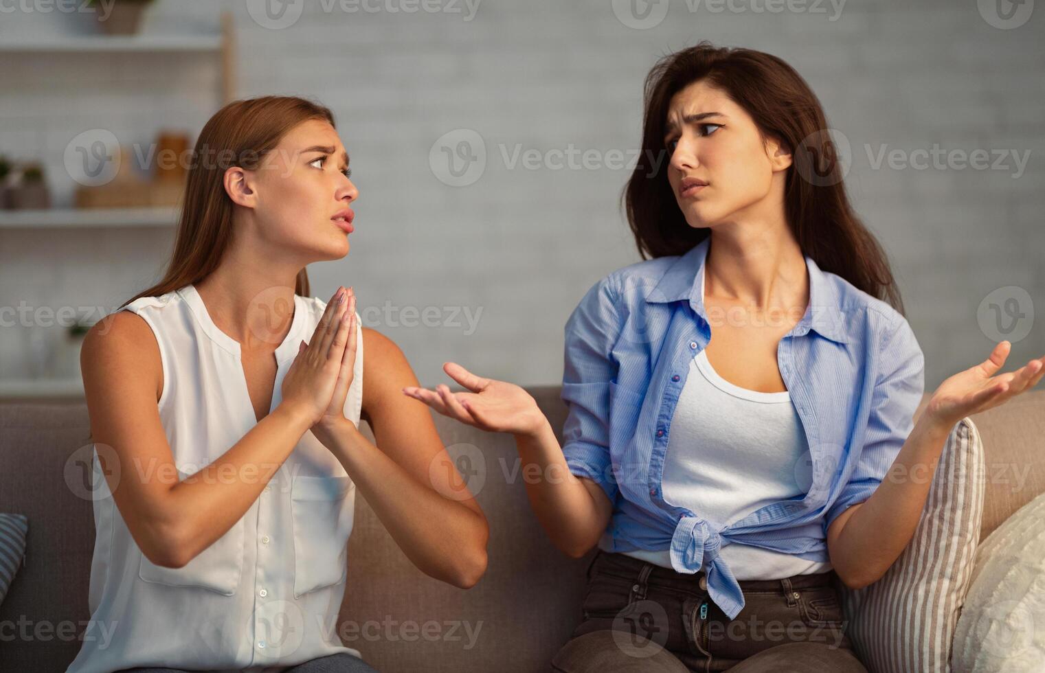 Bad Friendship. Girl Refusing To Do A Favor For Begging Friend Sitting On Sofa At Home. Selective Focus photo