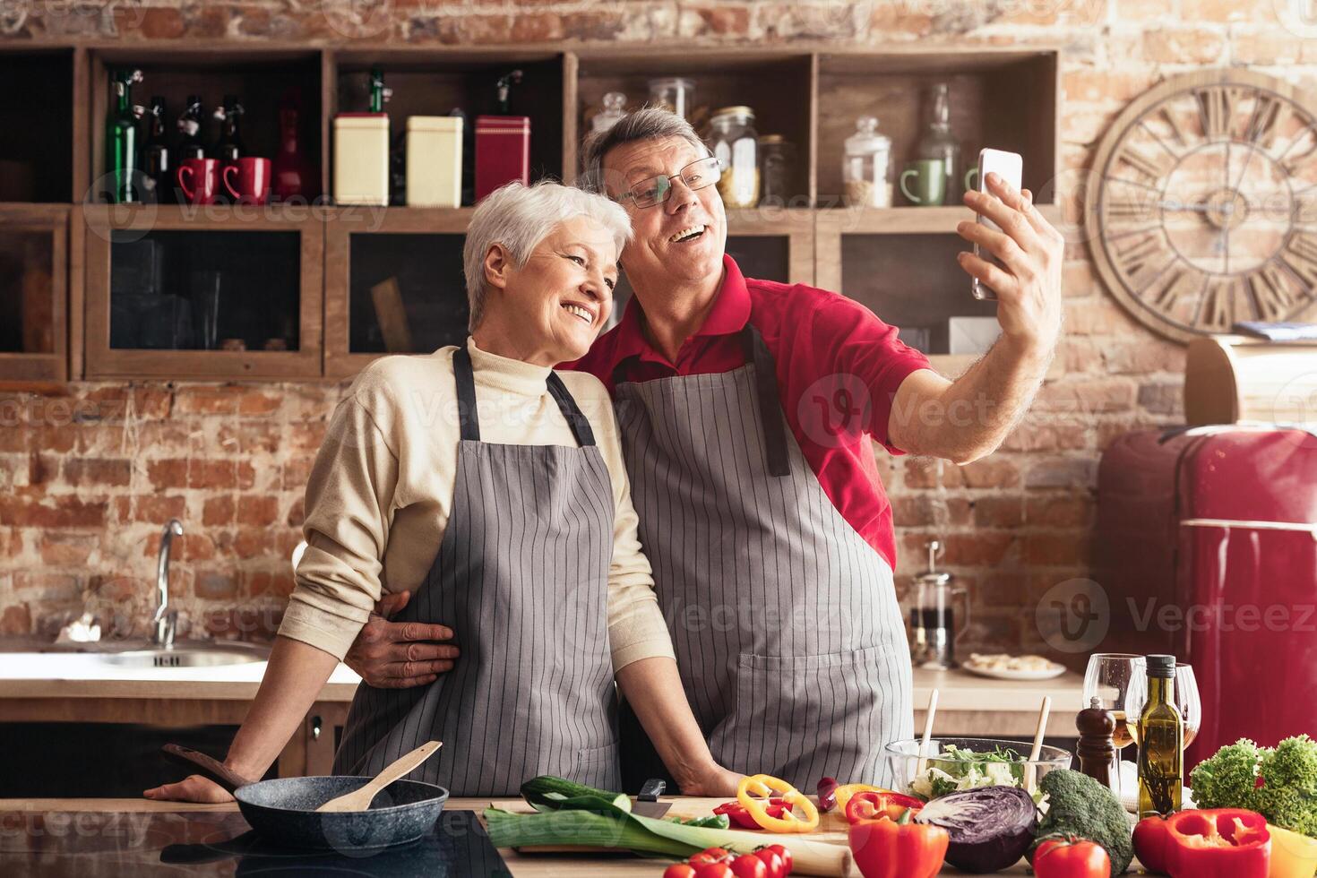 A happy older couple takes a selfie in a restaurant kitchen. The man is holding the phone, while the woman is standing beside him. They are both smiling and appear to be enjoying themselves. photo