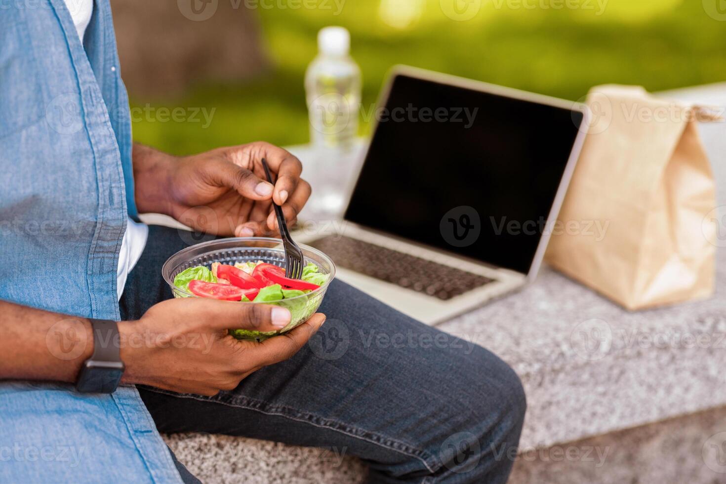 Lunch Break. Unrecognizable Afro Man Eating Fresh Vegetable Salad And Using Laptop With Black Screen On Bench In Park, Cropped Image, Closeup photo