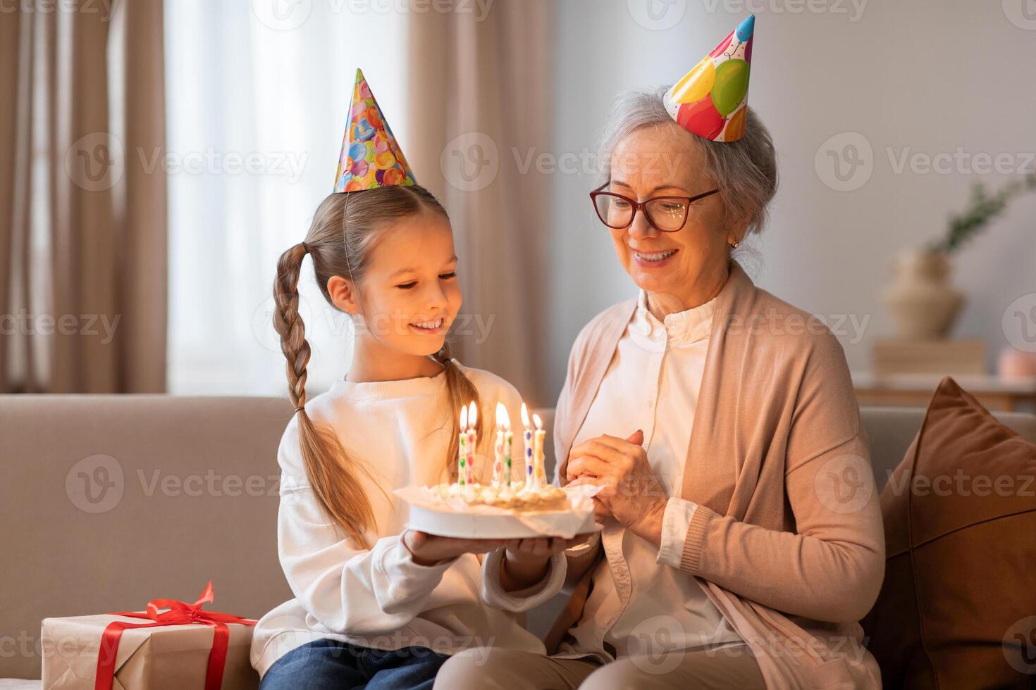 An older woman and a young girl sit on a couch, smiling at a birthday cake in front of them. The woman is lighting candles on the cake while the girl eagerly awaits a slice. photo