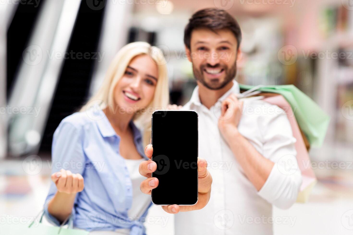 A man and woman, both smiling, stand in a shopping mall with bags, holding up a smartphone with black empty screen photo