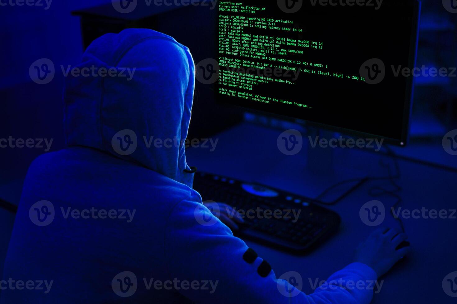 A person wearing a hooded sweatshirt sits in front of a computer with a dark background. Computer screen displays green code, indicating that the person is working on a programming or technical task. photo