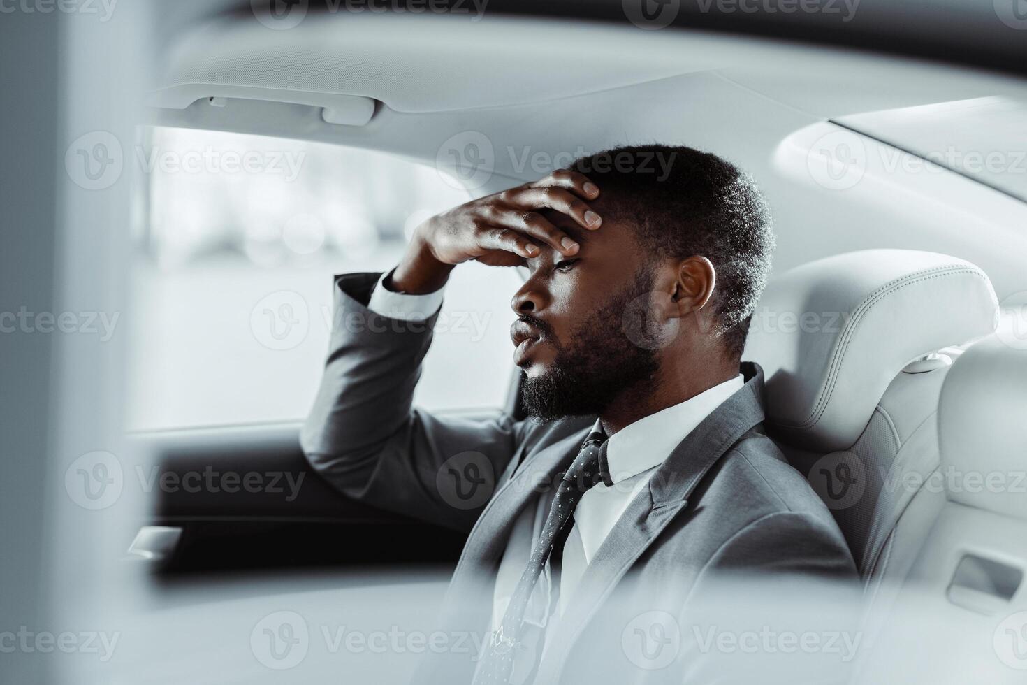A Black man in a grey suit sits in the back seat of a car. He looks down and has his hand on his forehead as if he is exhausted or stressed. The image is taken from inside the car photo