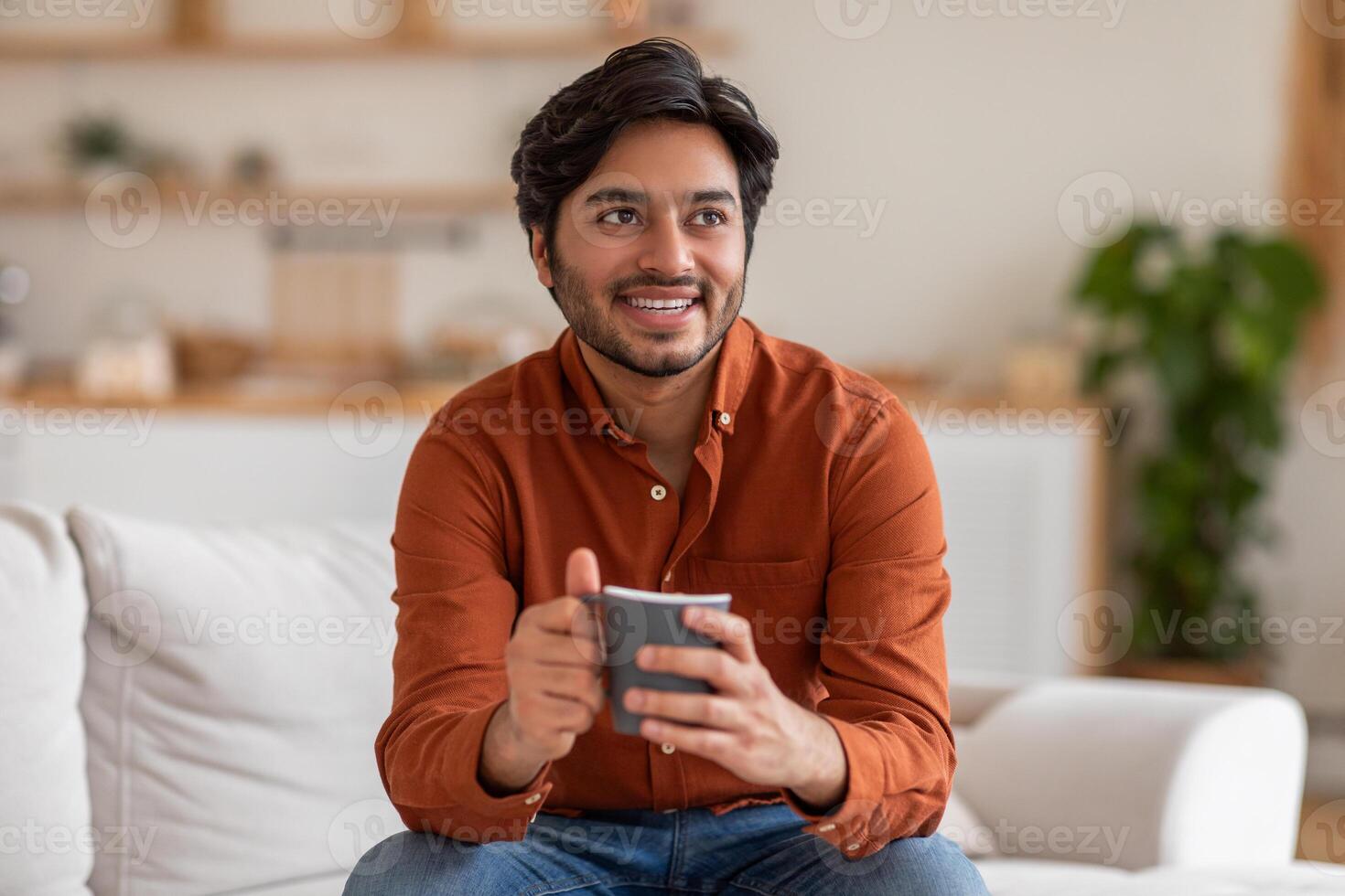 Arab man in an orange shirt sits on a white couch in his living room, smiling and holding a grey mug in his hands. He looks off to the side, likely deep in thought or enjoying a moment of peace photo