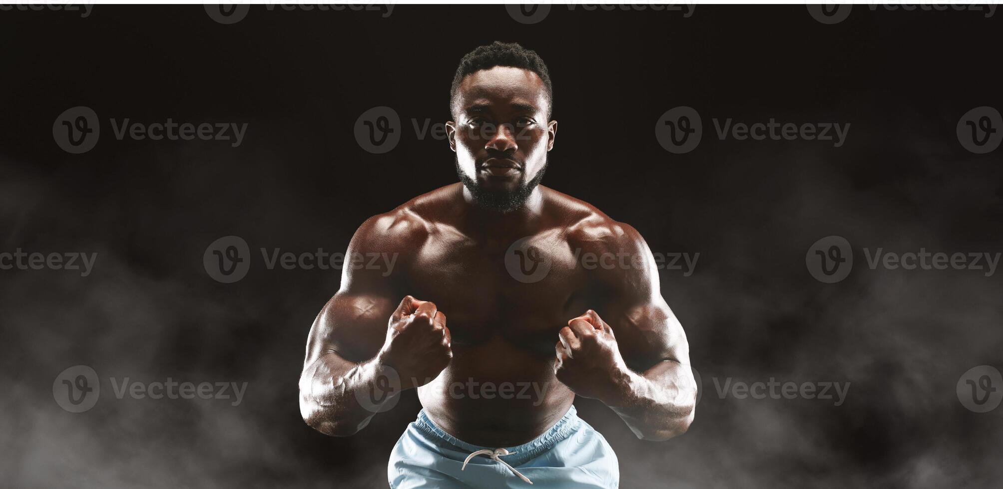 A muscular man with a determined expression poses against a dark background with smoke swirling around him. photo