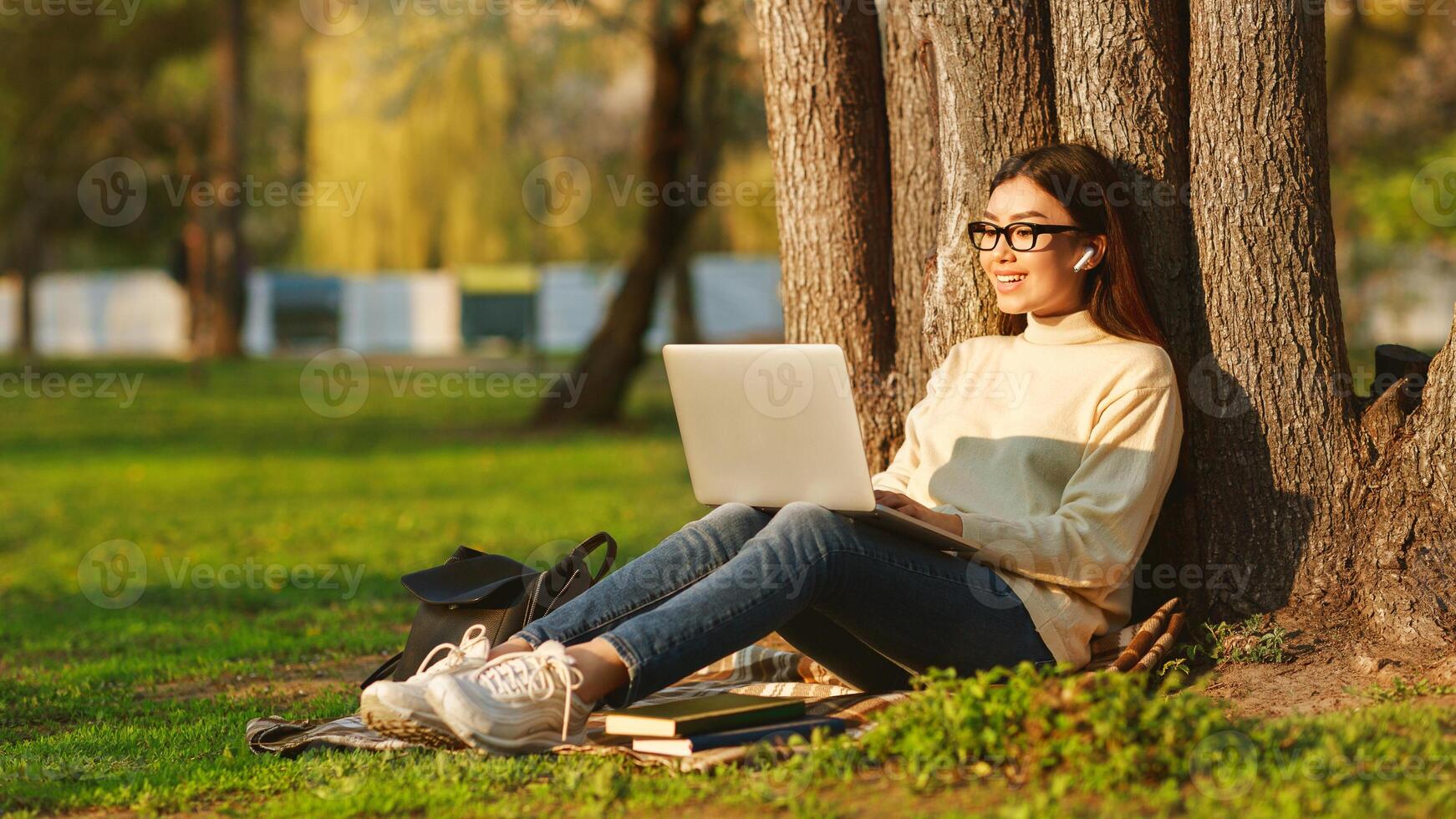 Asian young woman is studying on her laptop while sitting on the grass in a park. The atmosphere is serene and productive, capturing the balance of nature and technology. photo