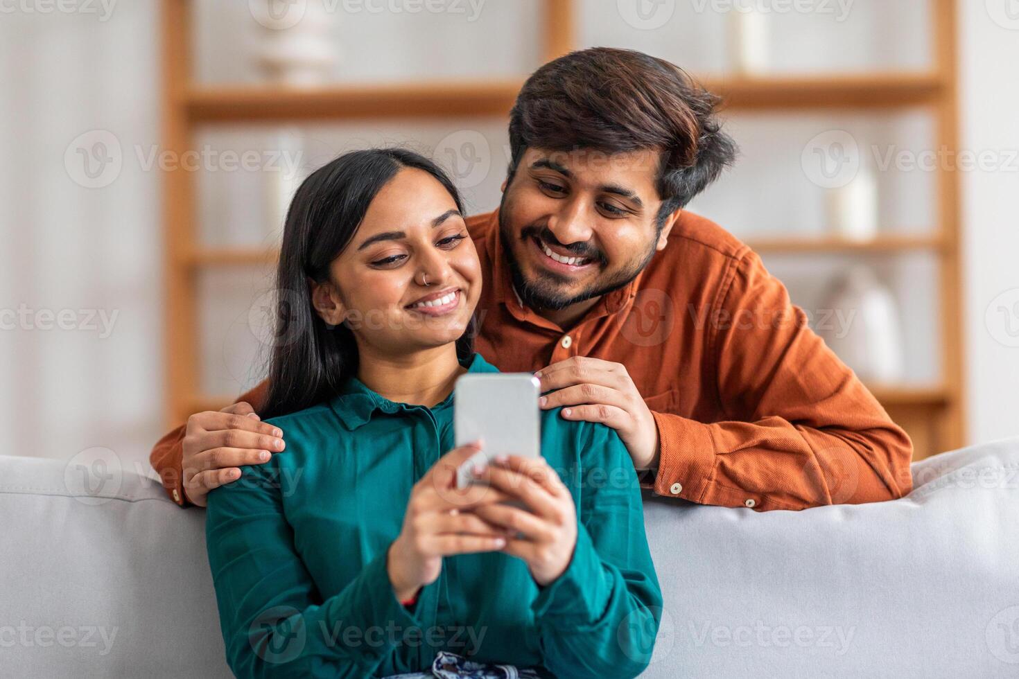 Indian man and woman are sitting together on a couch, both focused on a cell phone in the mans hand. They appear engrossed in the screen, possibly watching something or browsing social media. photo