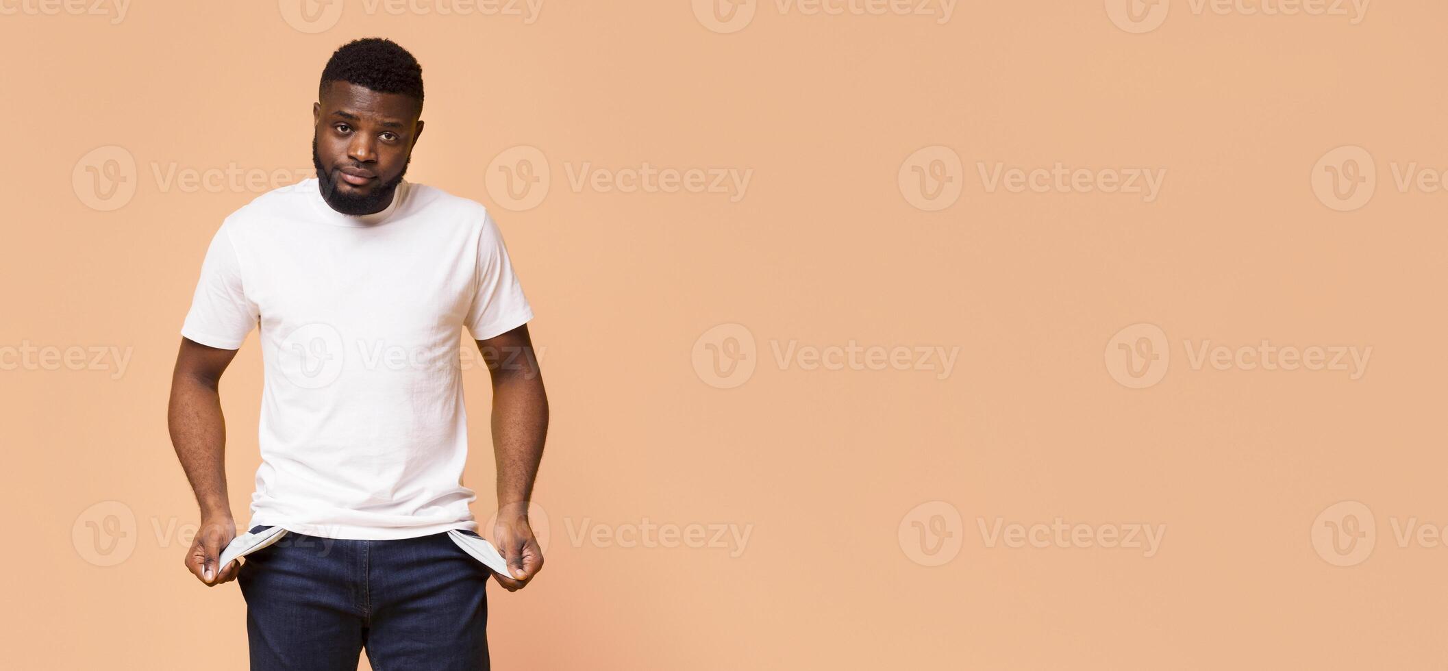 A young black man with a beard is standing against a beige backdrop. He is wearing a white t-shirt and blue jeans and is checking his pockets, which appear to be empty, copy space photo