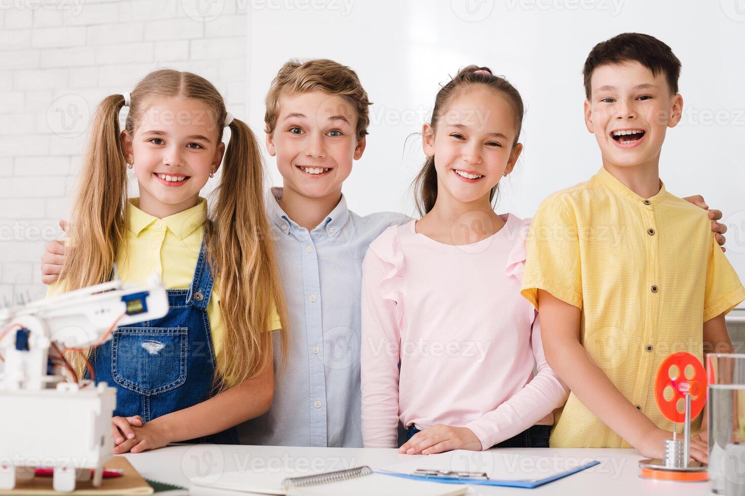 Four young students, two boys and two girls, are smiling and working on a science experiment in a classroom. They are all wearing casual clothing and are standing around a table with a robot photo