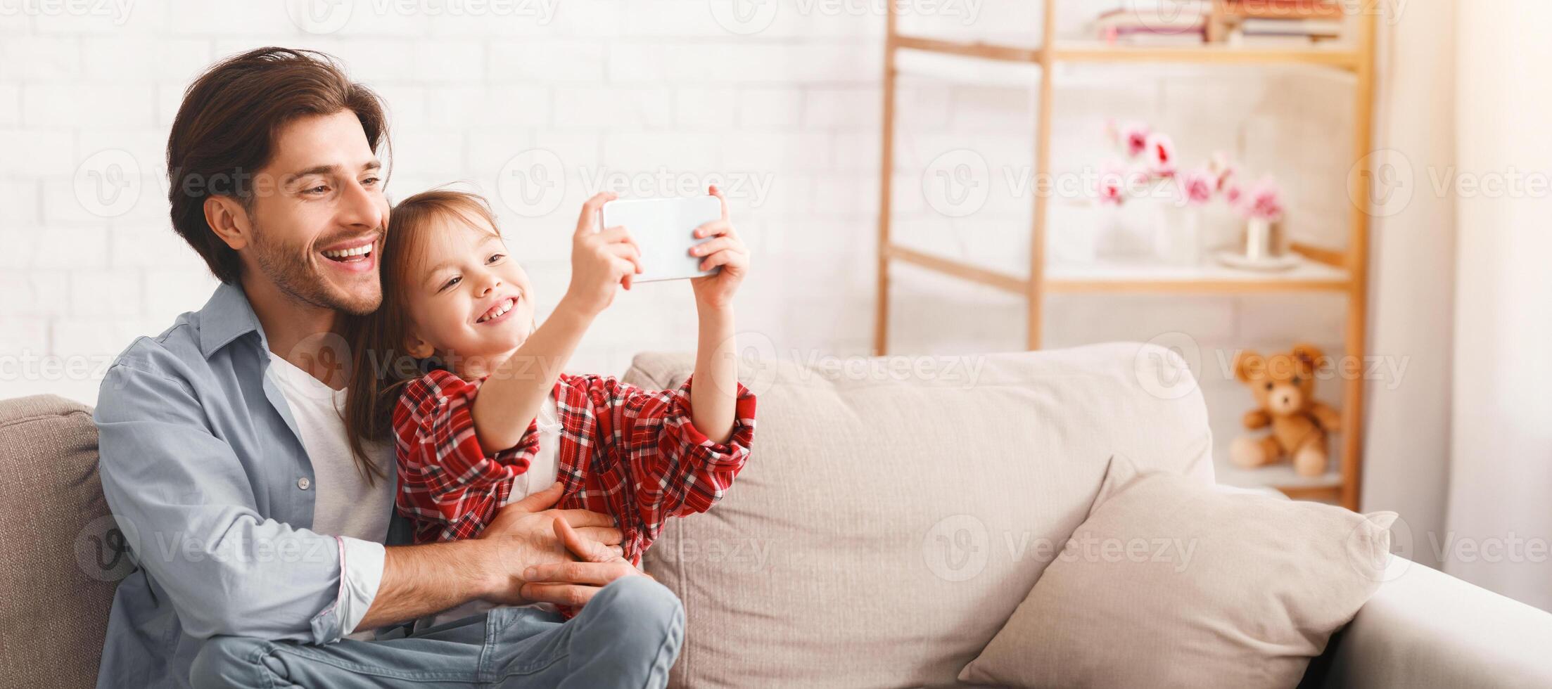 Preschool girl taking selfie with her cheerful father, sitting on sofa at home, copy space photo