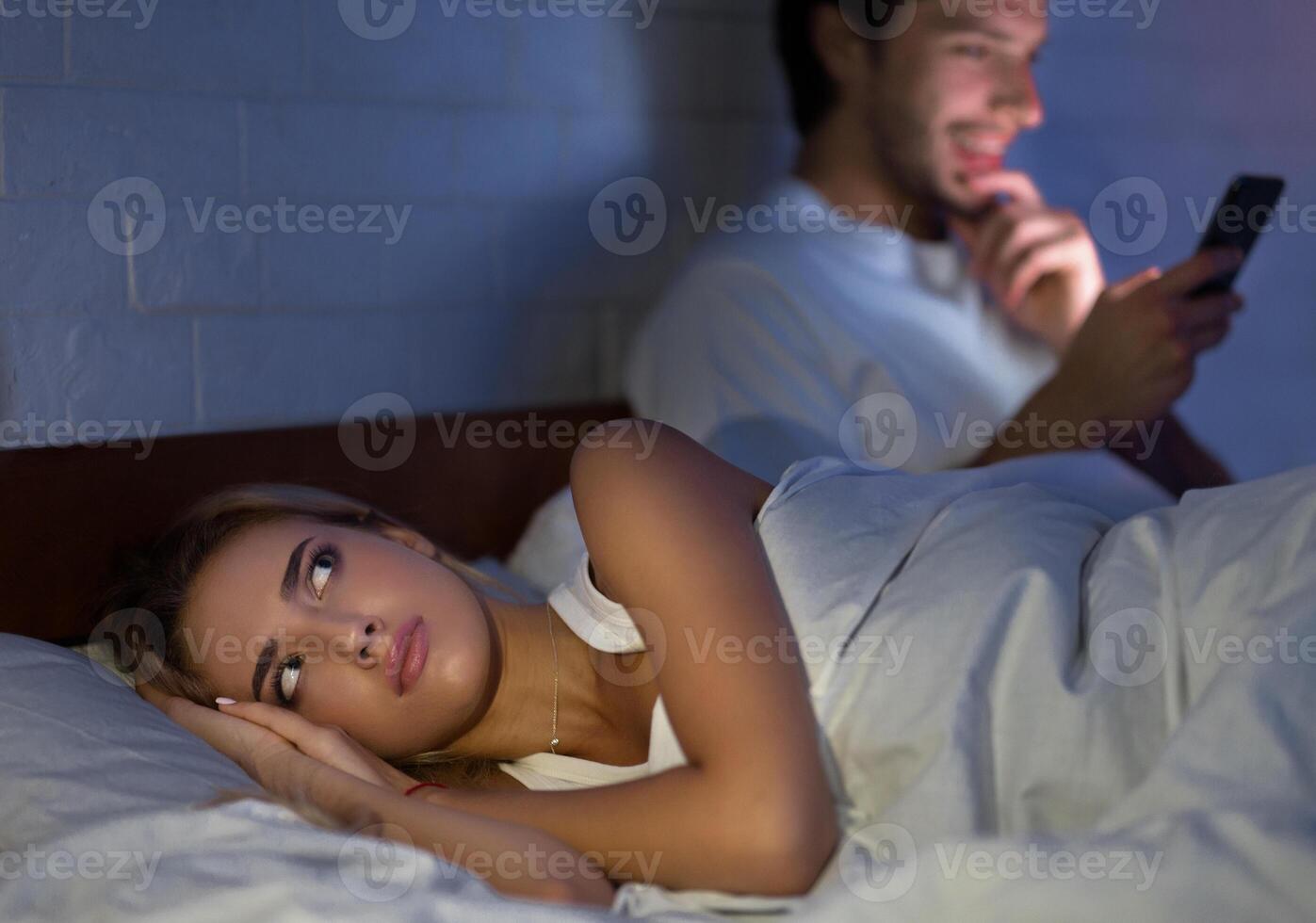 Suspecting Infidelity. Man Texting On Mobile Phone Lying In Bed With Wife At Night. Low Light, Selective Focus photo