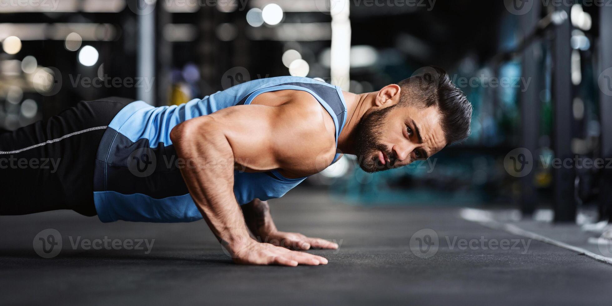Young man workout in fitness club. Profile portrait of strong caucasian guy making plank or push ups exercise, training indoors, empty space photo