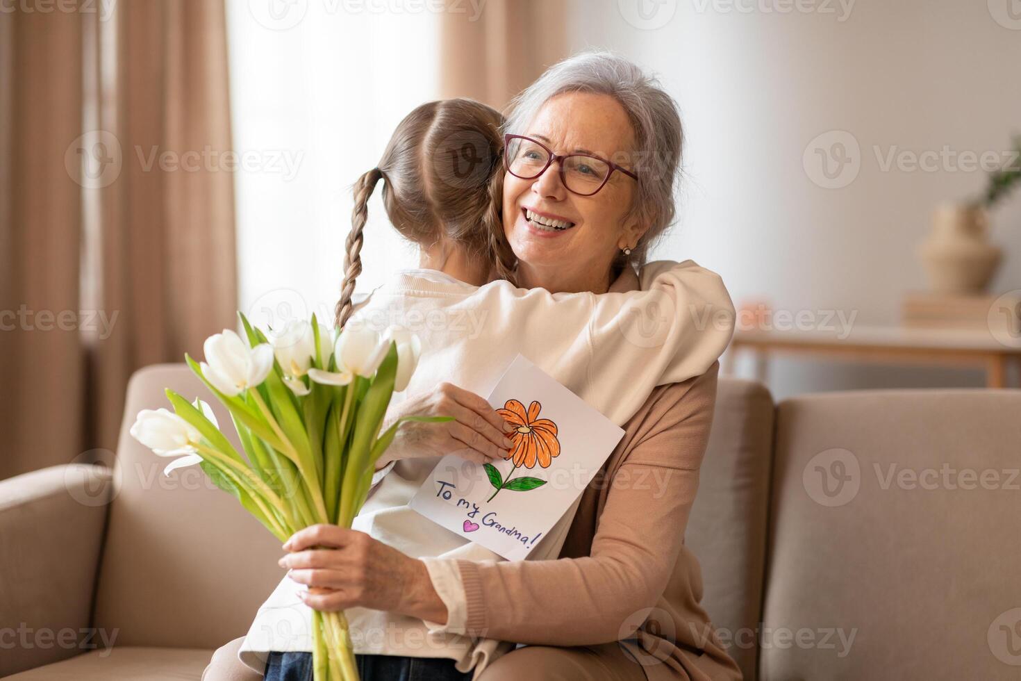 A heartwarming scene unfolds as a young girl with braided hair embraces her smiling grandmother, presenting her with a bouquet of fresh white flowers and a colorful handmade card. photo