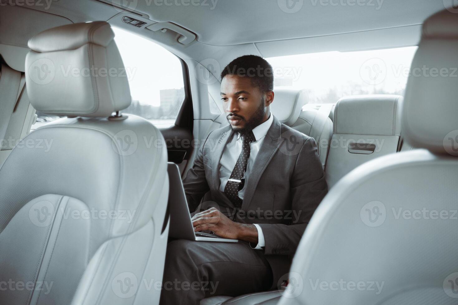 Black businessman in a suit is sitting in the back seat of a car, working on his laptop computer. He is focused on his work, typing on the keyboard. The cars interior is clean and modern photo