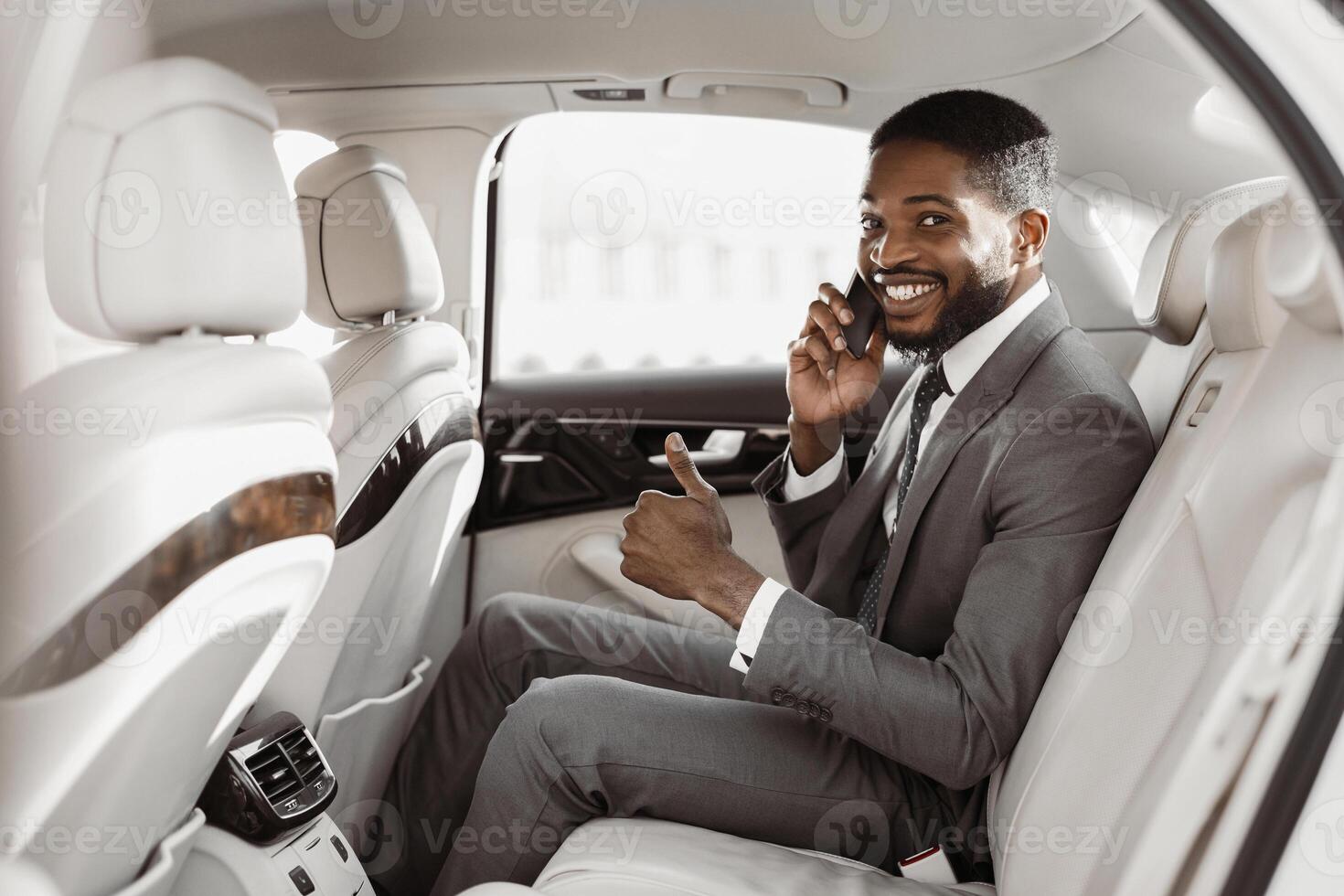 African American successful businessman, dressed in a suit, sits in the backseat of a car. He is talking on his phone, smiling, and giving a thumbs up. The car is clean and luxurious photo
