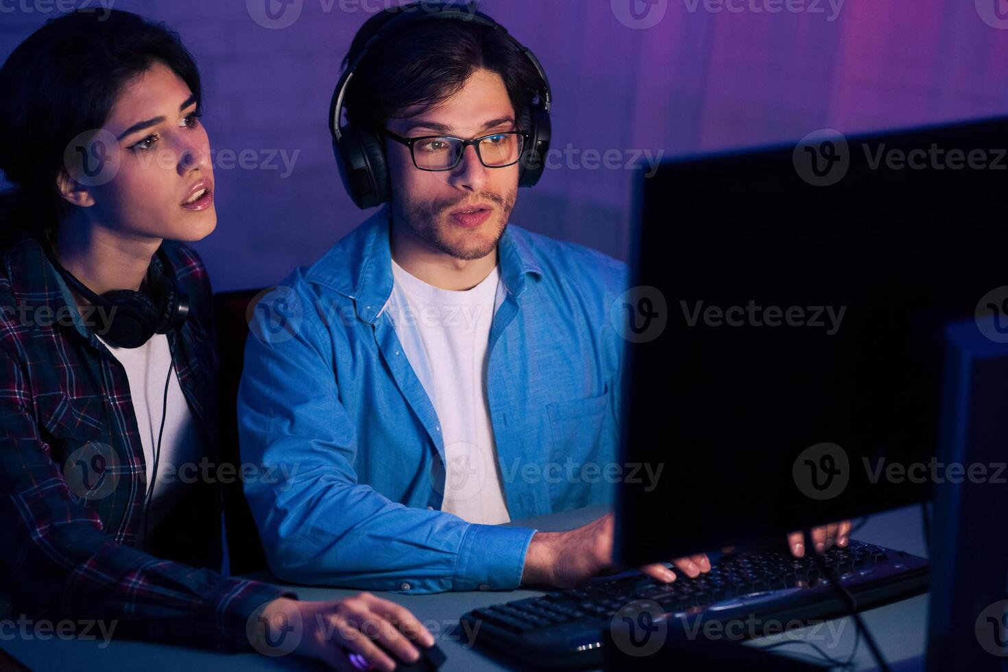 A young man and woman are sitting at a desk in front of a desktop computer, both wearing headphones and looking intently at the screen. photo
