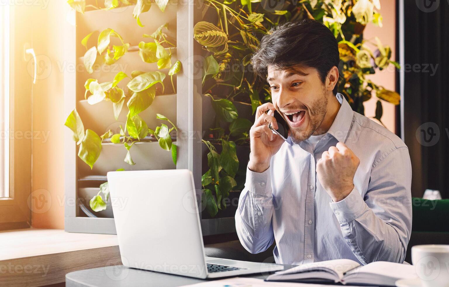 Unbelievable success. Excited millennial businessman in cafe with open laptop, looking at computer screen and talking on phone, empty space photo