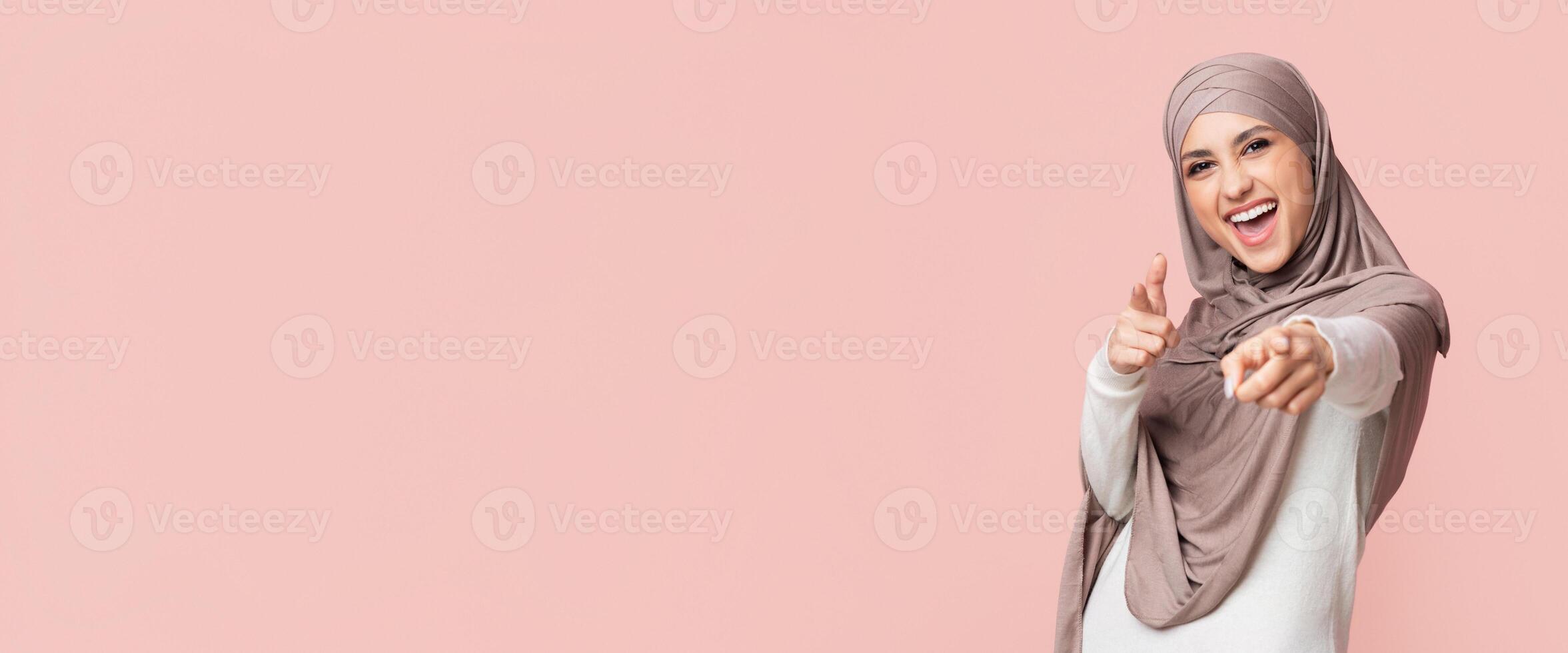 Gotcha. Positive Muslim Girl In Hijab Pointing Fingers At Camera, Posing On Pink Background, Empty Space photo