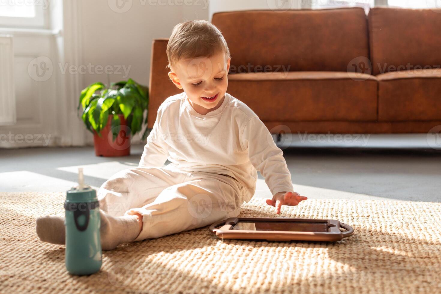 A young boy is seated on the floor, engrossed in playing with a tablet device. His focus is on the screen as he interacts with the digital content photo
