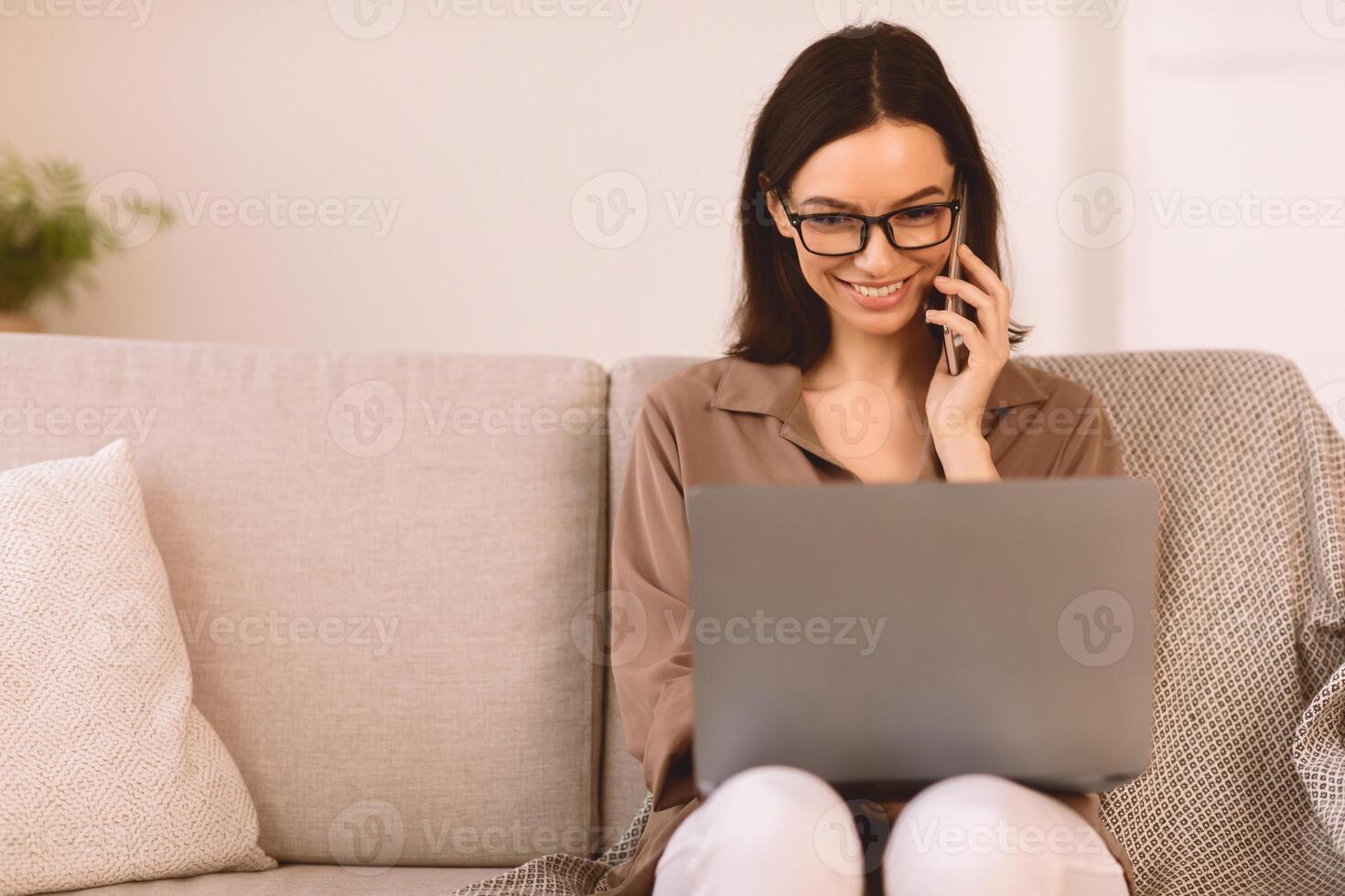 Corporate Communication. Smiling girl talking on mobile phone working on laptop at home, sitting on sofa, copyspace photo