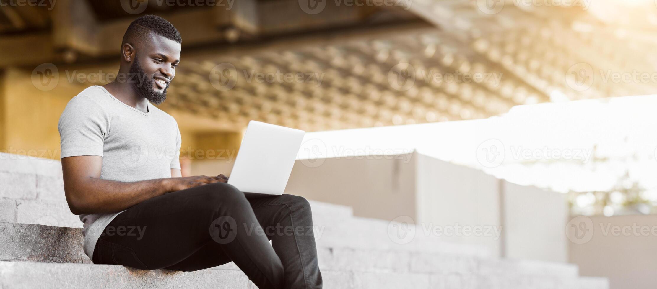 Smiling african american guy coding on his laptop and using city wifi outdoor, panorama, copy space photo