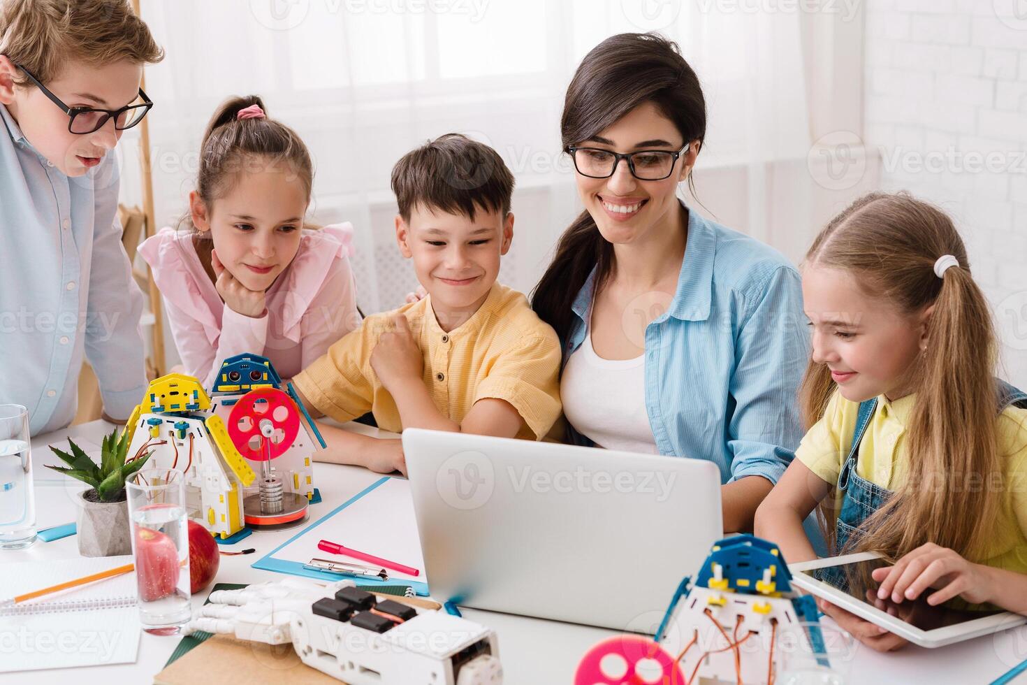 Elementary school students are working on a robotics project in their classroom. The students are gathered around a laptop and are looking at the screen intently. Their teacher is sitting beside them photo