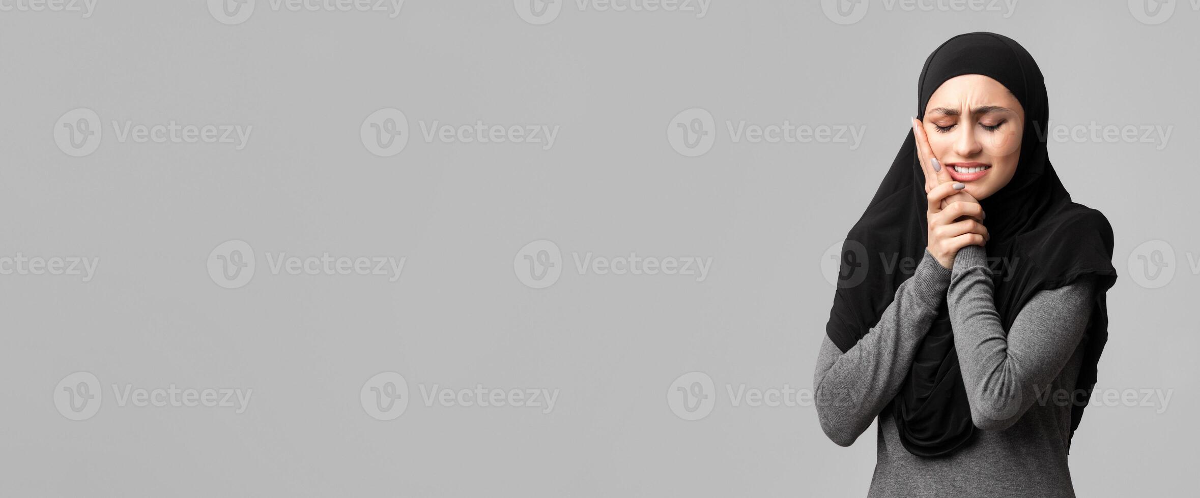 Toothache. Stressed arabic girl in hijab touching her cheek, suffering from hypersensitive teeth, standing over gray background in studio, free space photo