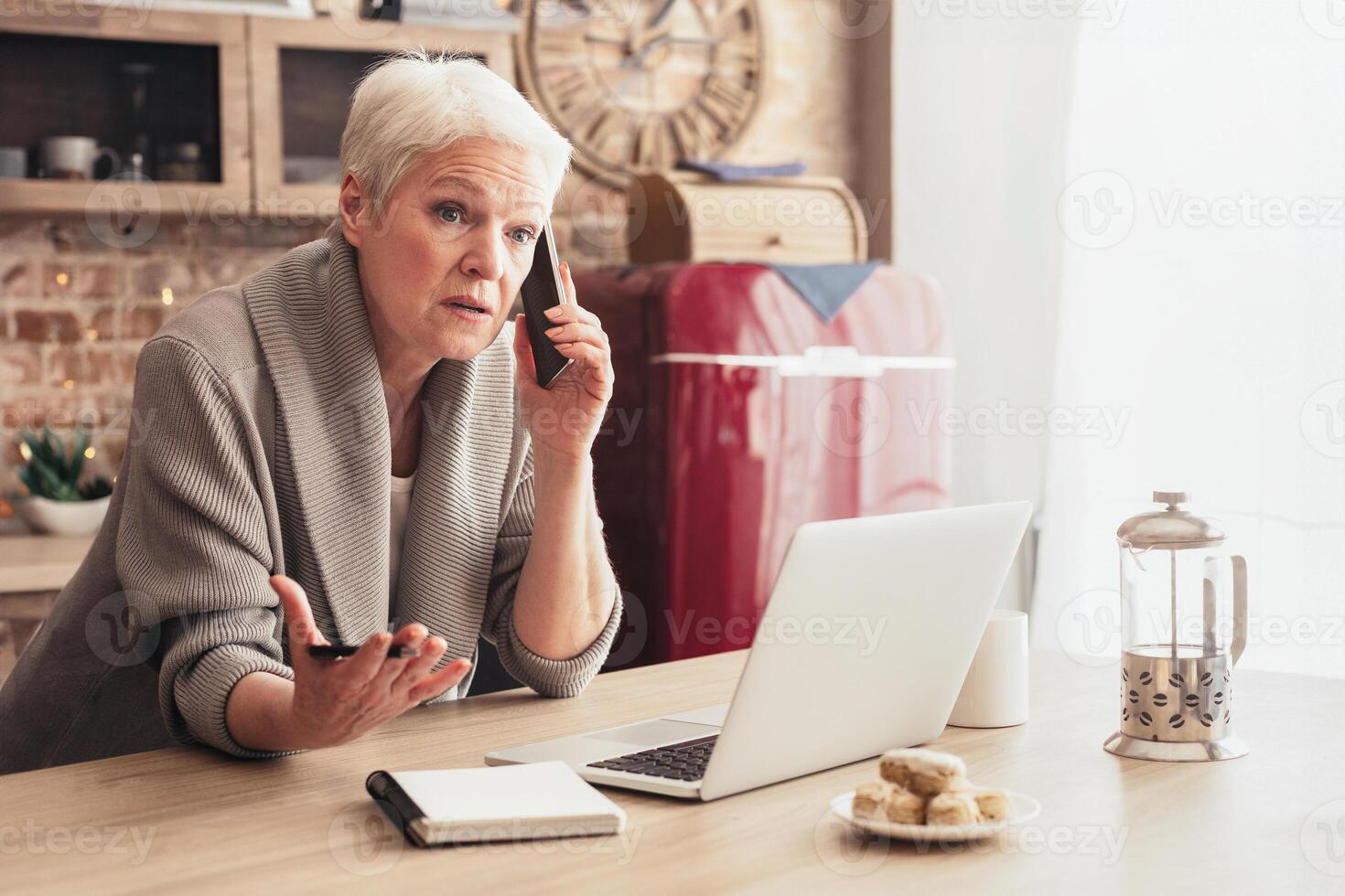 A senior woman with white hair sits at a kitchen table talking on a mobile phone while gesturing with her other hand. She is sitting in front of a laptop computer and a notebook. photo