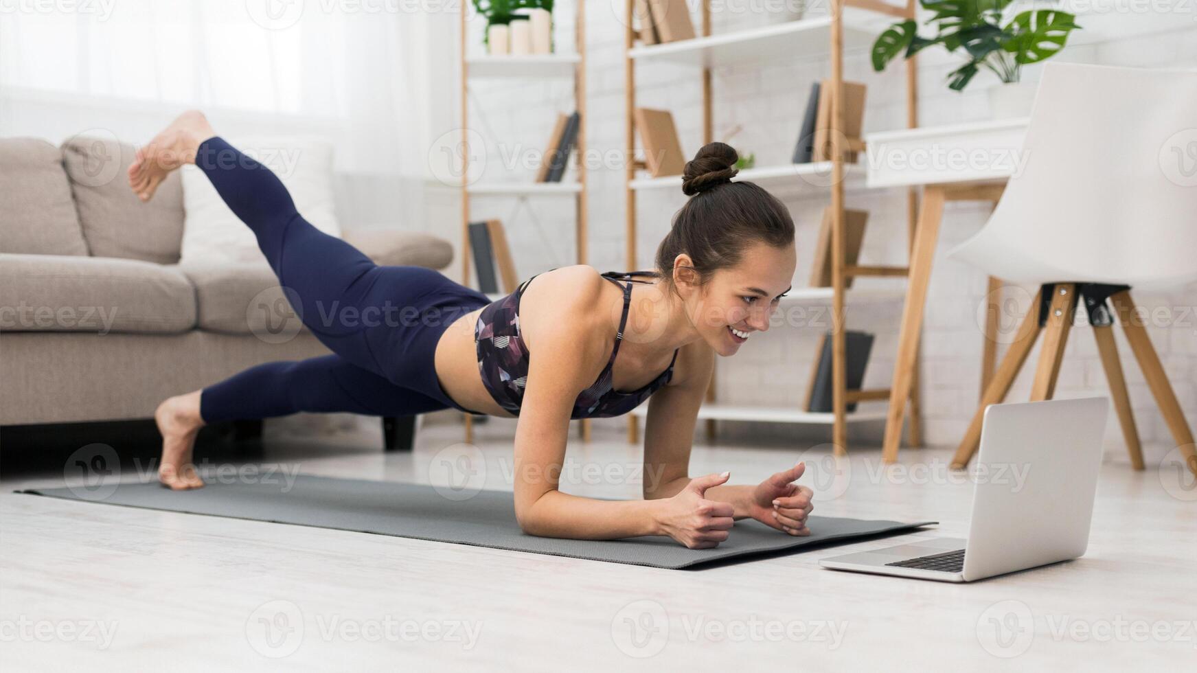 A woman in athletic wear is doing a plank exercise on a yoga mat while watching a laptop. She is in her living room with a couch and bookshelf in the background. photo