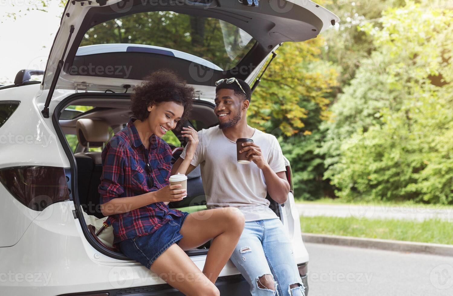 Happy afrocouple sitting in open car back, having stop off, drinking coffee. Romantic traveling concept photo