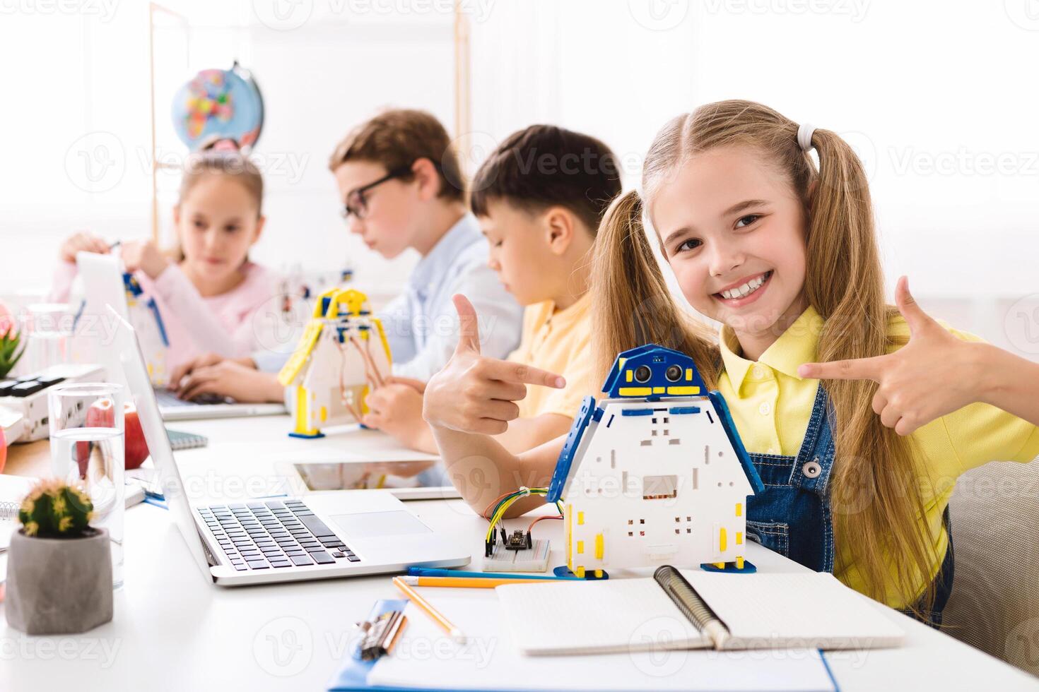 A young girl smiles proudly as she points to a robot she built in a STEM classroom setting. The image shows other students working on similar projects and a laptop with a robotic arm on the desk. photo