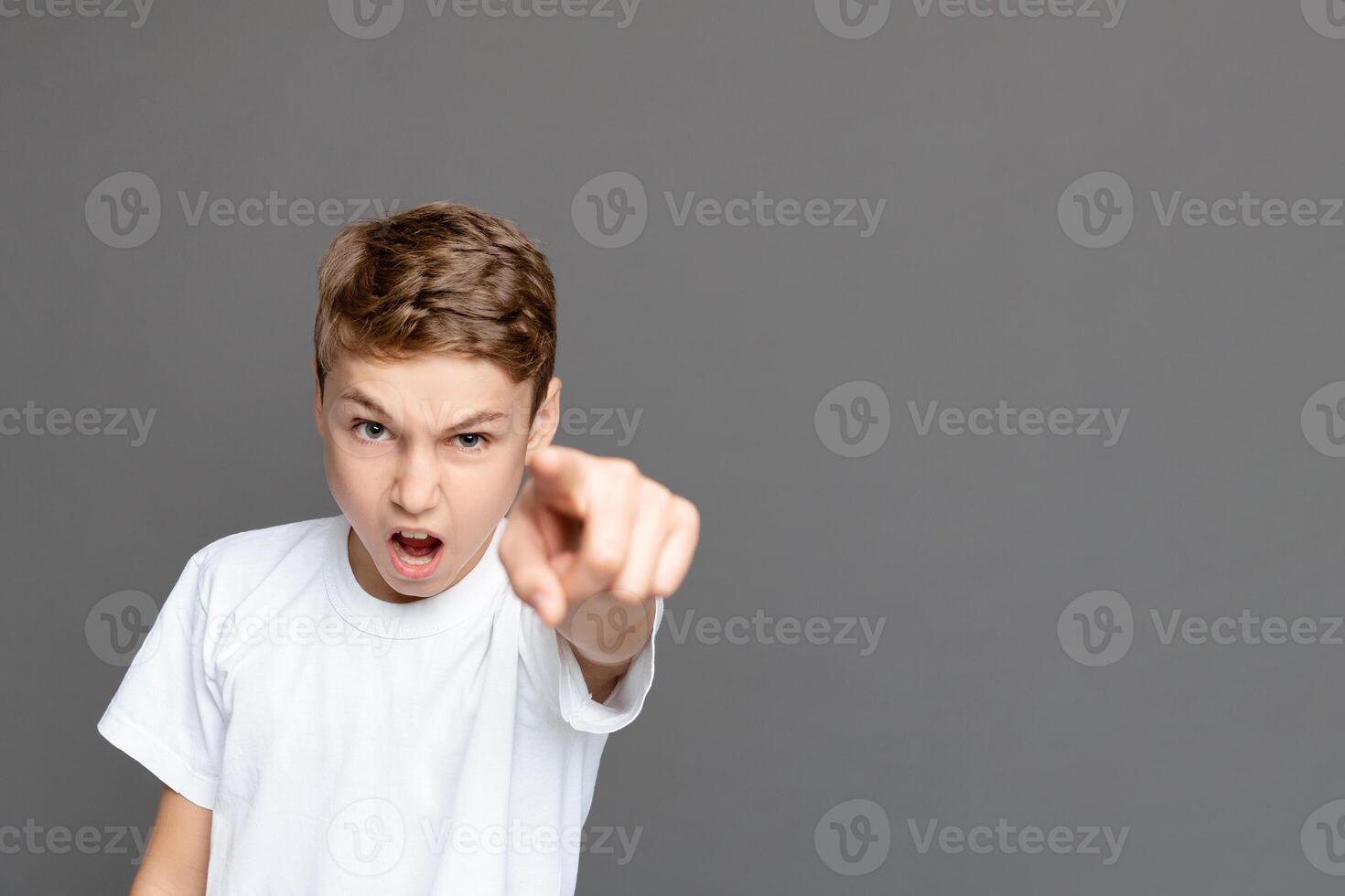 A young boy with an assertive expression points directly at the camera, conveying directness or accusation, standing isolated against a grey background photo
