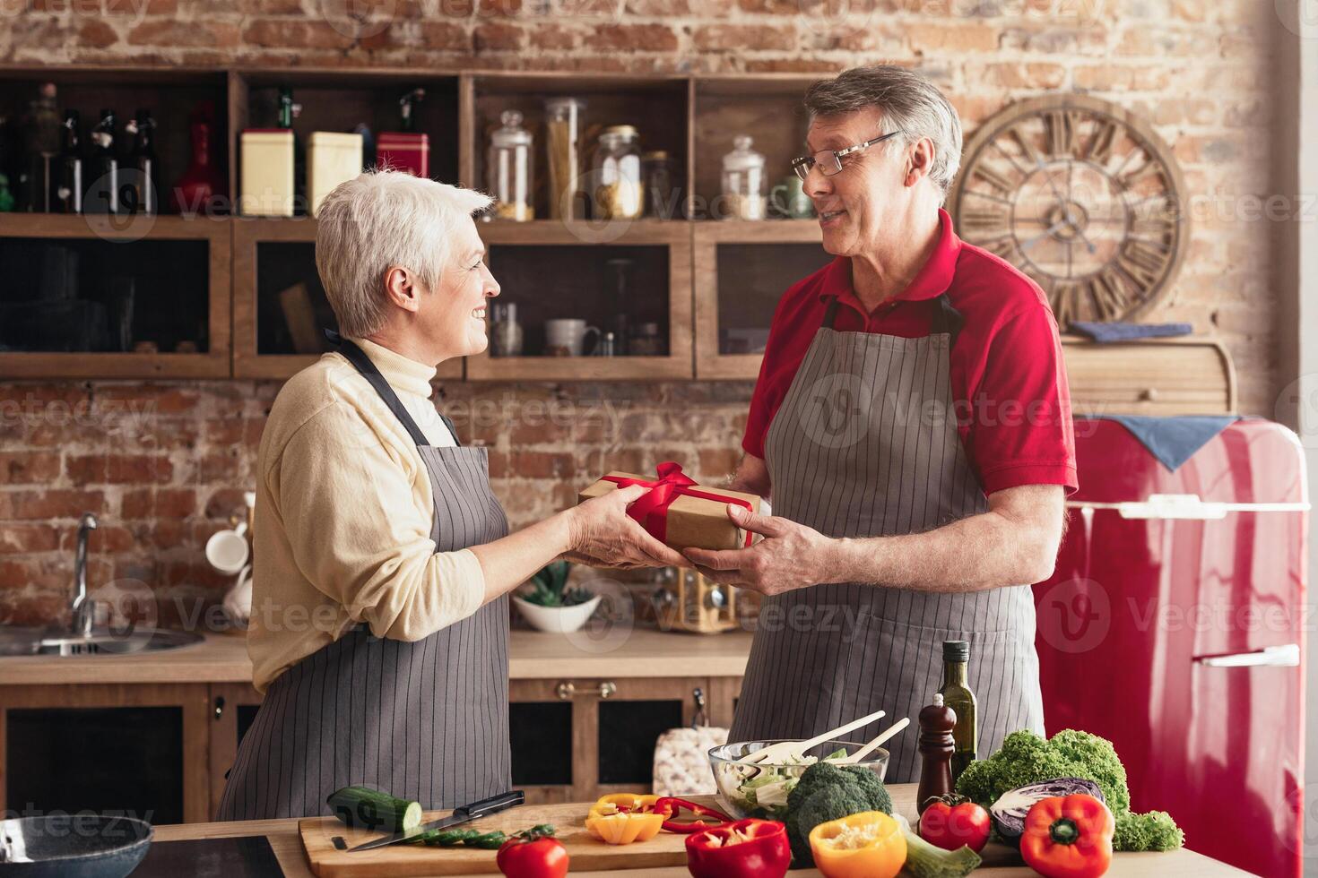 A senior couple stands in a kitchen, exchanging a wrapped gift. The woman is smiling and holding the gift out to the man, who is smiling back at her. There is a red refrigerator in the background photo