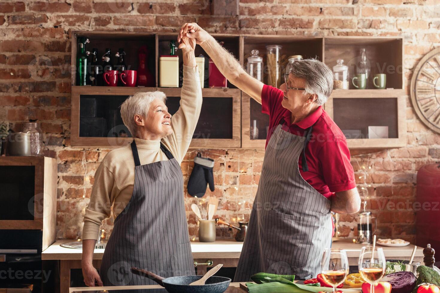 A senior couple in grey striped aprons happily dance in a kitchen with a brick wall and wooden cabinets. They are holding hands and smiling as they twirl. The kitchen counter is full of vegetables photo