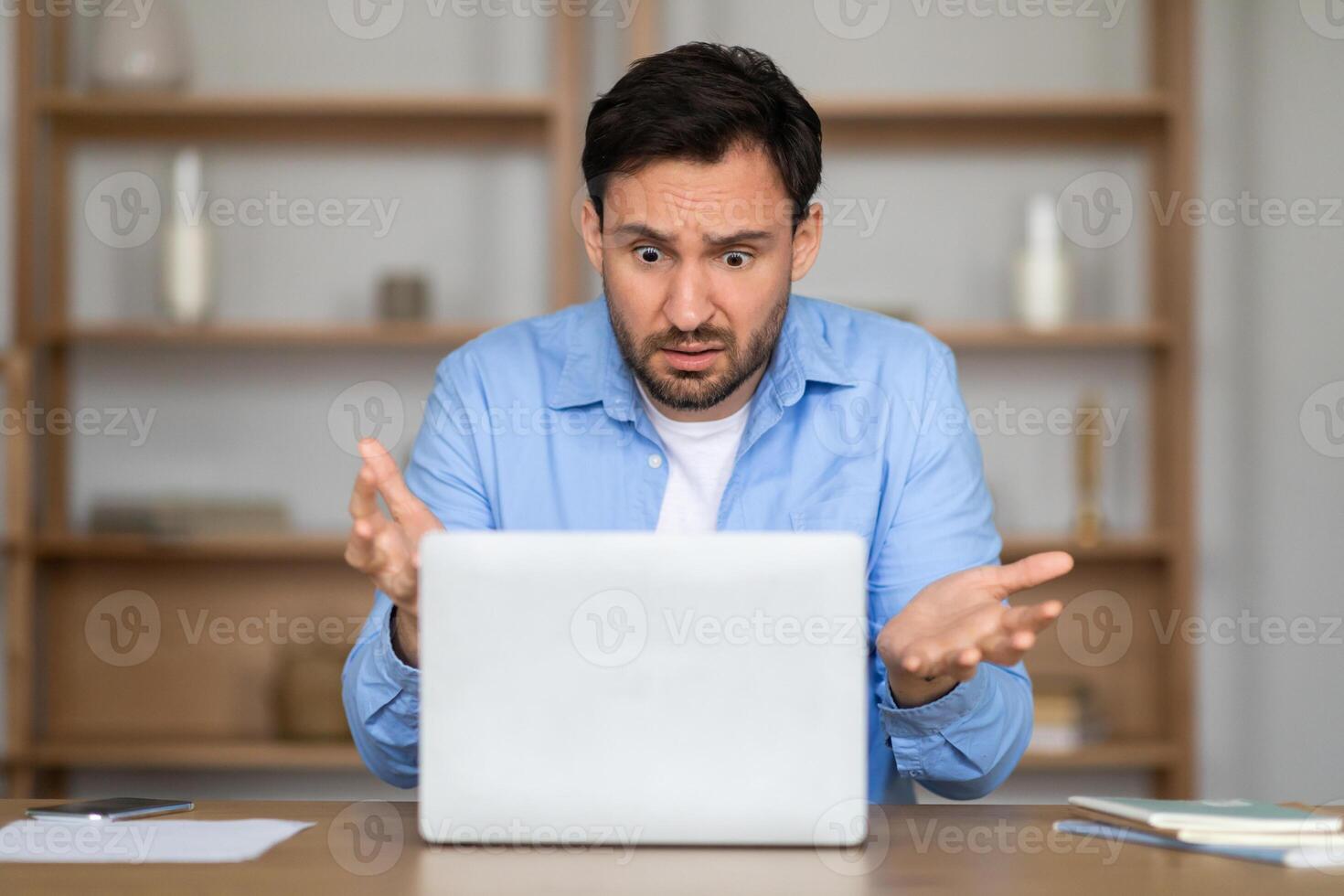 A man sits at a desk with an expression of confusion or surprise as he stares at his laptop screen, with papers scattered around him suggesting he is in the midst of work photo