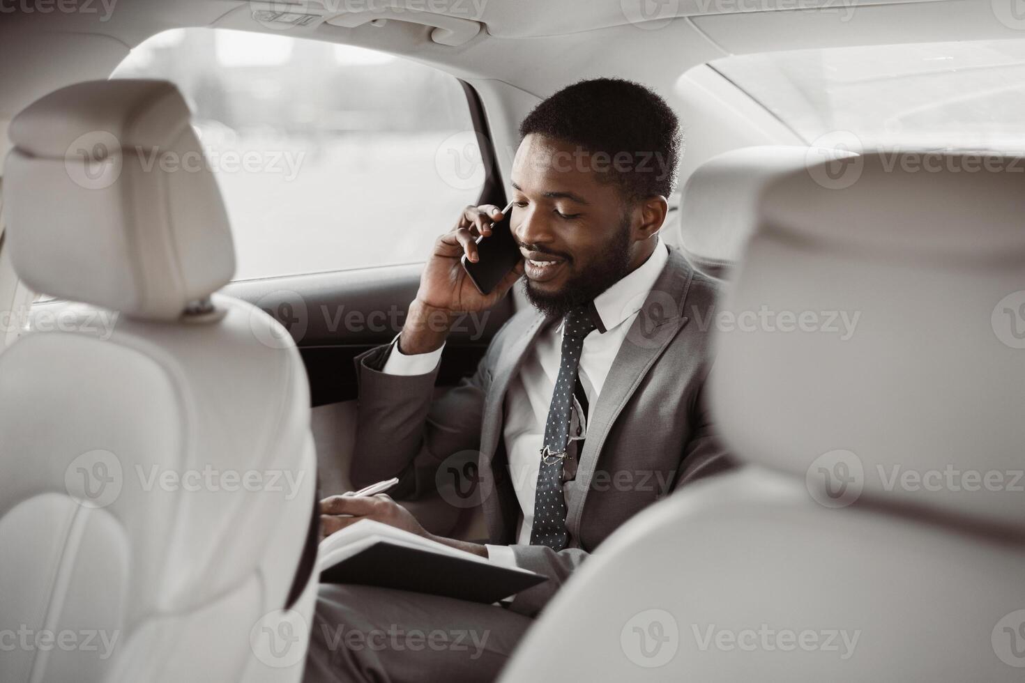 African American businessman in a suit sits in the backseat of a car. He is talking on his phone and taking notes in a notebook. He appears to be engaged in a business conversation while commuting. photo
