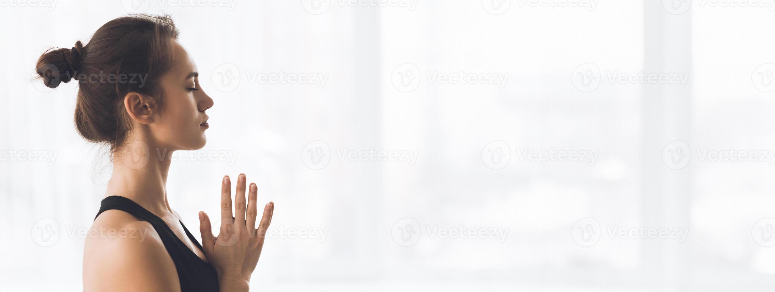 A young woman stands in a peaceful pose with hands together, focusing on her breath in a well-lit room, copy space photo