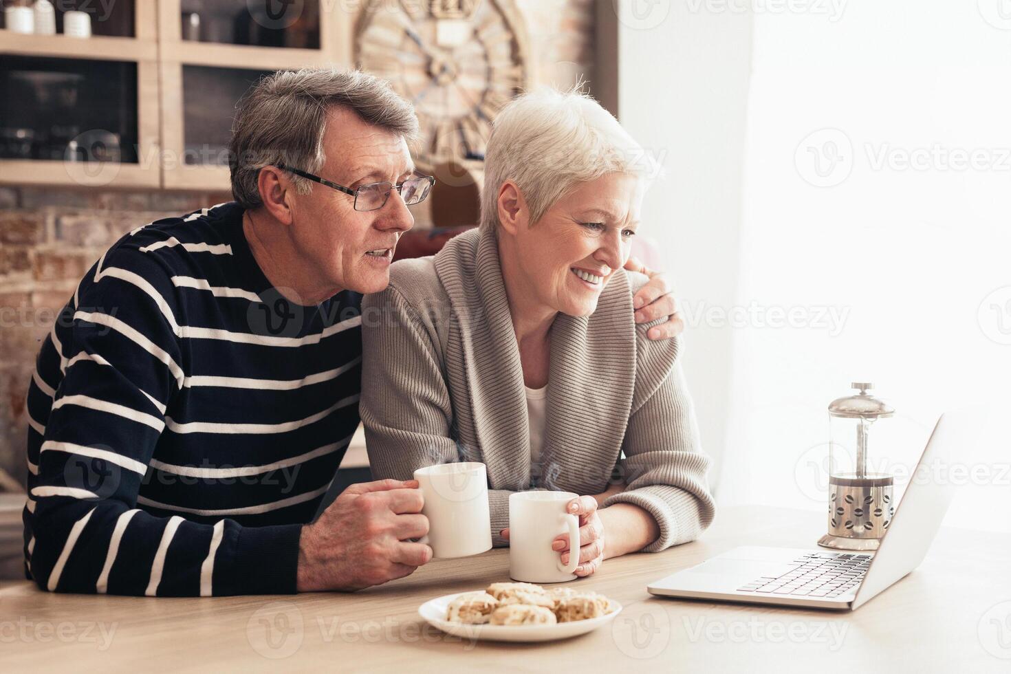 A senior couple is sitting in their kitchen, enjoying coffee and conversation while using a laptop. The woman is smiling at the screen, while the man is looking at her photo